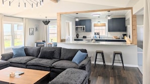 Un salon moderne avec un canapé d’angle, une table basse en bois et une cuisine dotée de meubles sombres et d’un grand îlot central.