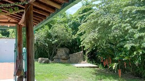 A covered outdoor area with a wooden structure, a paved entrance, and a lush garden with various plants and a rock feature.