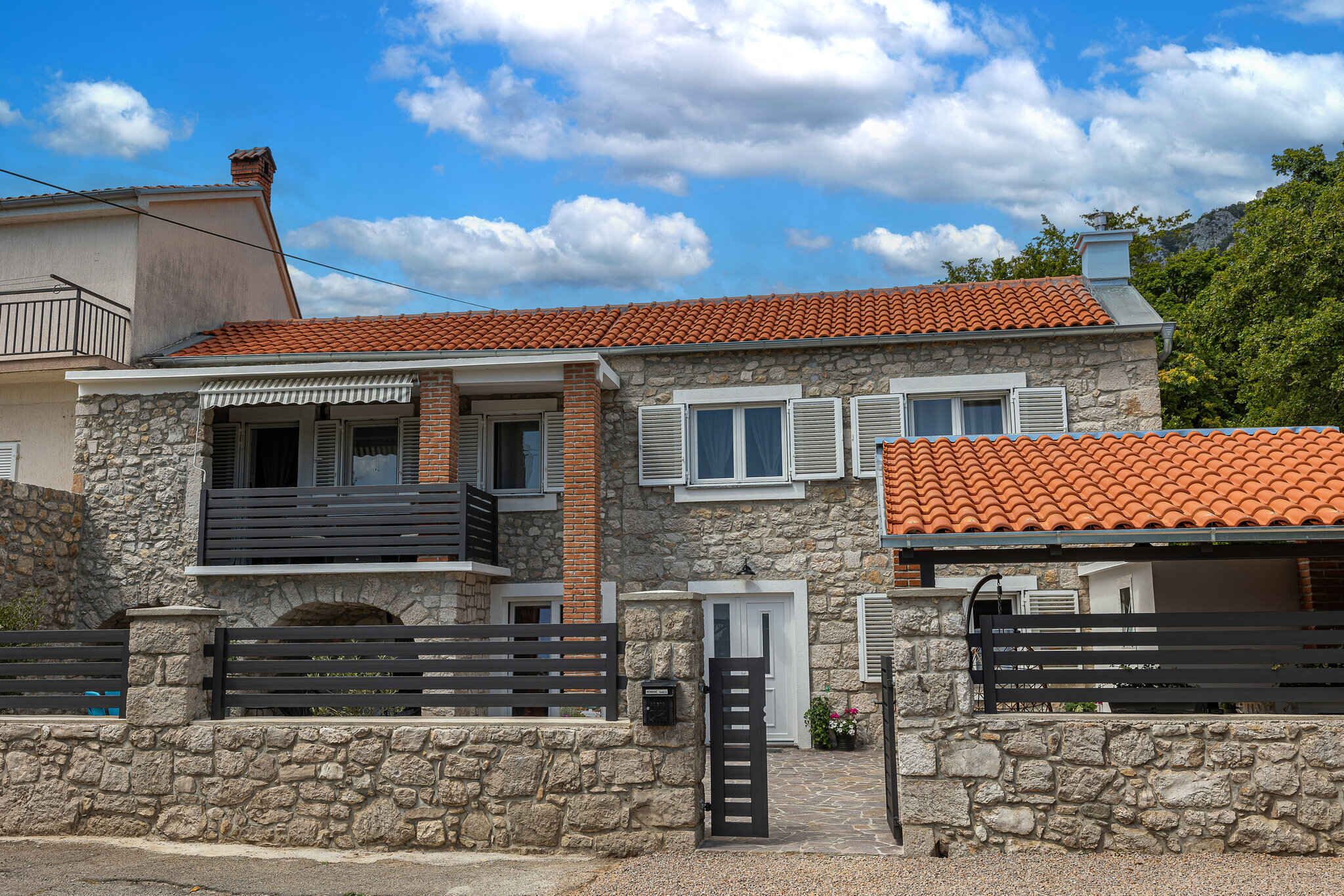 Une maison en pierre à deux étages, avec un toit recouvert de tuiles rouges, un mur en pierre et un portail noir.