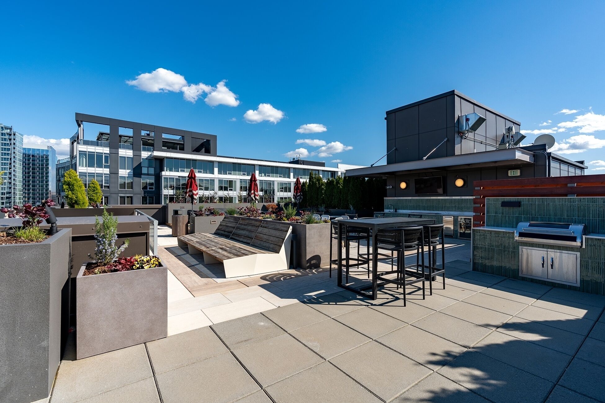 Una terraza en la azotea con mobiliario exterior, zona de barbacoa y vistas a edificios modernos.