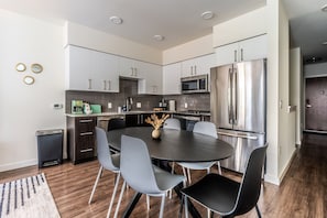 A modern kitchen with white cabinets, stainless steel appliances, and a dining area with a round table and chairs. - 2BR TH | Gym, Balcony | Bellevue Parkside M (Bellevue)