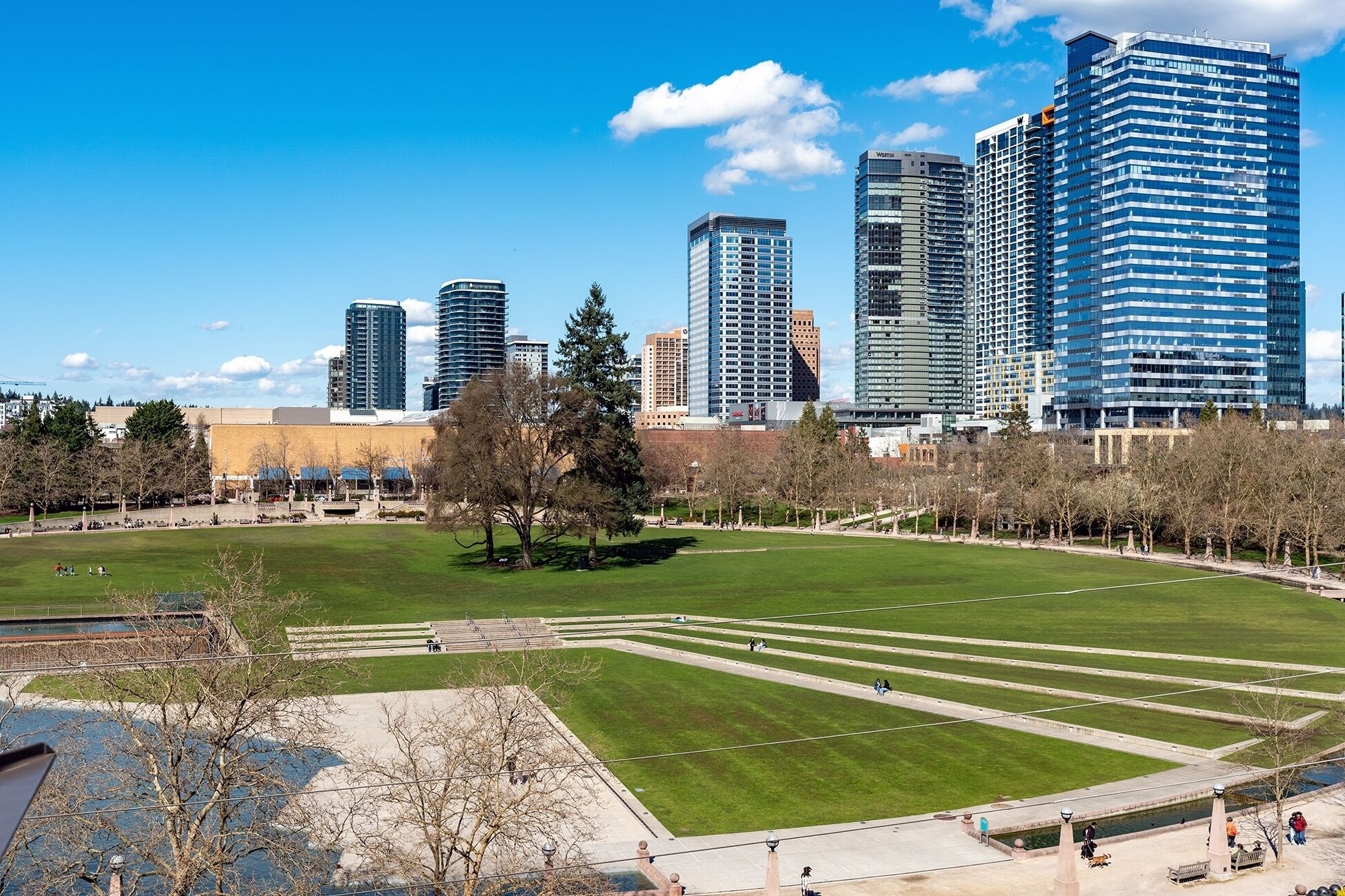 A cityscape with modern high-rise buildings, a large open park with pathways, and a body of water in the foreground.