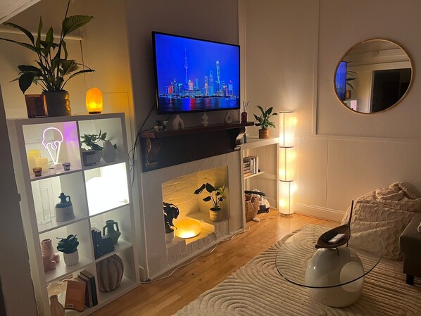 Un salon avec un téléviseur diffusant une vue sur la ville, une cheminée, des étagères ornées d’objets décoratifs et un grand miroir rond accroché au mur.
