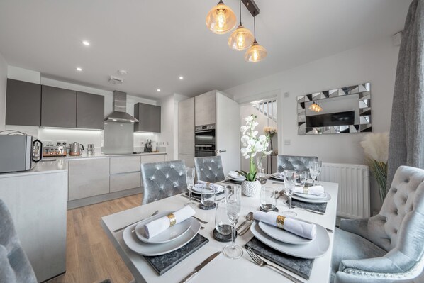 A modern dining area with a white table set for four, grey upholstered chairs, and a grey sofa. - 3 bedroom house in Bricket Wood , St Albans with Free Parking and EV Charger (Bricket Wood)