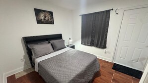 A bedroom with a bed, a nightstand, a wall-mounted picture, a door, and a window with curtains. - Room, cozy and well-lit (Tampa)