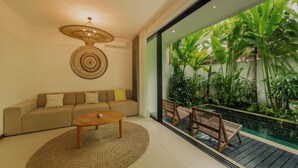 A modern living room with a sofa, coffee table, and a view of a tropical garden through a glass door. - Premier Umalas Seminyak (Keroboken Kelod)