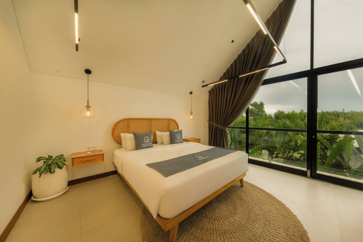 A modern bedroom with a large bed, bedside tables, a potted plant, and a view through large windows.