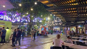 Un espace de restauration en plein air, avec des tables et des chaises, entouré d’arbres et illuminé de guirlandes lumineuses.