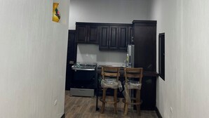Un pasillo estrecho con una lámpara moderna en el techo, sillas de madera, una zona de cocina con armarios oscuros y un refrigerador.