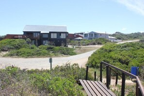 A modern building with a flat roof and large windows, surrounded by greenery and a sandy path.