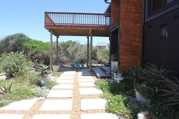 A covered outdoor area with a wooden deck, potted plants, and a pathway leading to a building.
