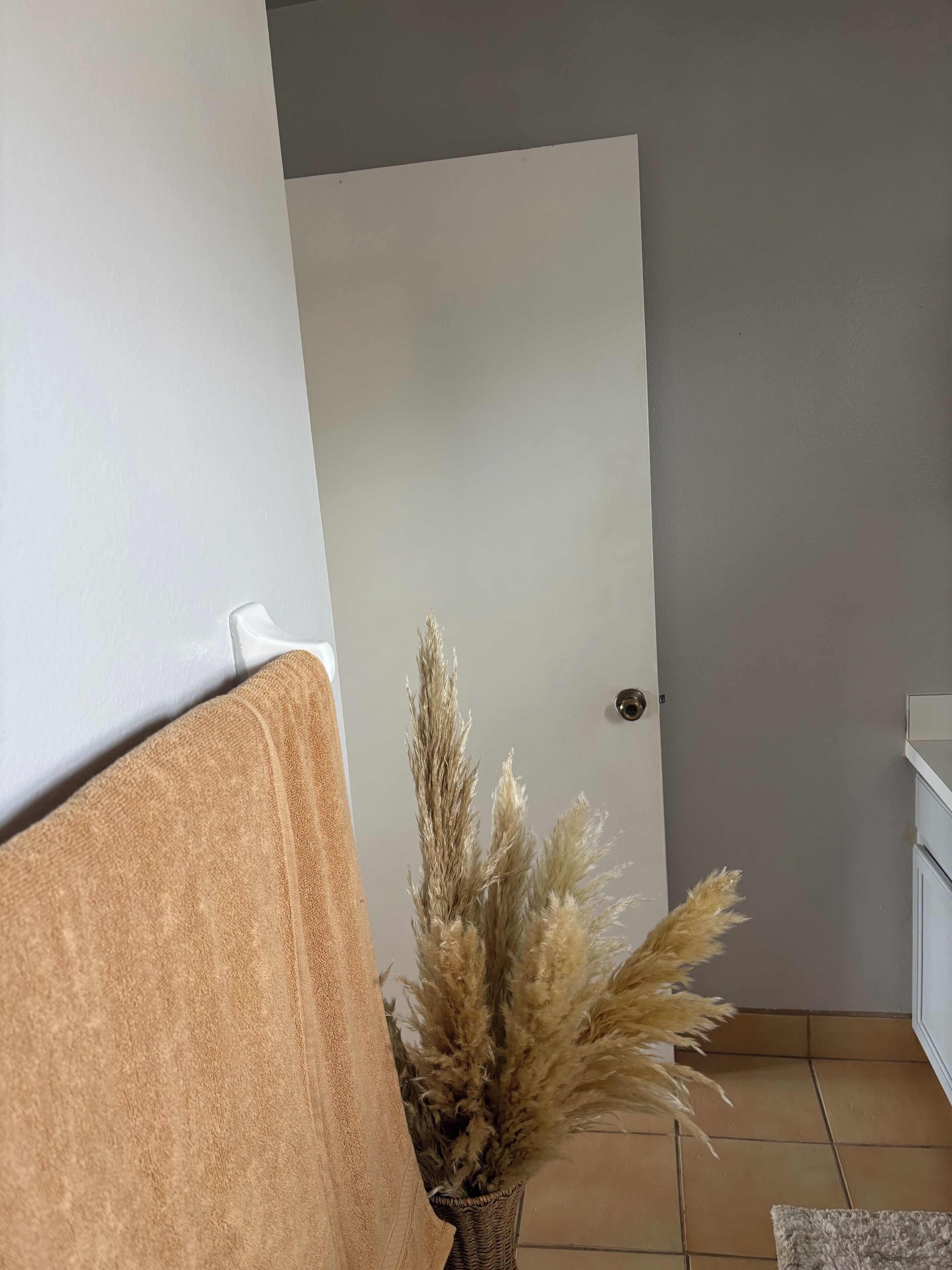 Une pièce avec une porte blanche, une serviette beige posée sur une chaise et un vase contenant des plantes séchées.