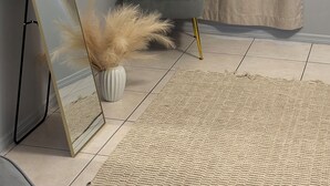 Una stanza con una poltrona grigio scuro, uno specchio a figura intera, un vaso con pampas essiccate e un tappeto decorativo su un pavimento piastrellato.