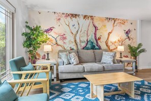 A living room with a grey sofa, teal armchairs, a wooden coffee table, and a colorful mural of trees and birds. - 2BR 1.5BA Gorgeous Condo Near Hospitals - Shopping (Saint Andrews)