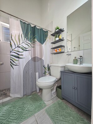 A bathroom with a shower curtain, toilet, sink, and green rugs. - 2-bedroom brand new central townhome in soothing Portmore with air conditioning (Portmore)