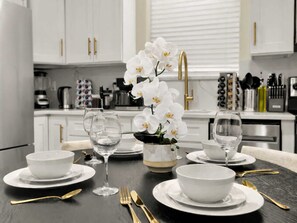 Una mesa de comedor servida con platos blancos, cuencos, copas de vino y cubiertos dorados.