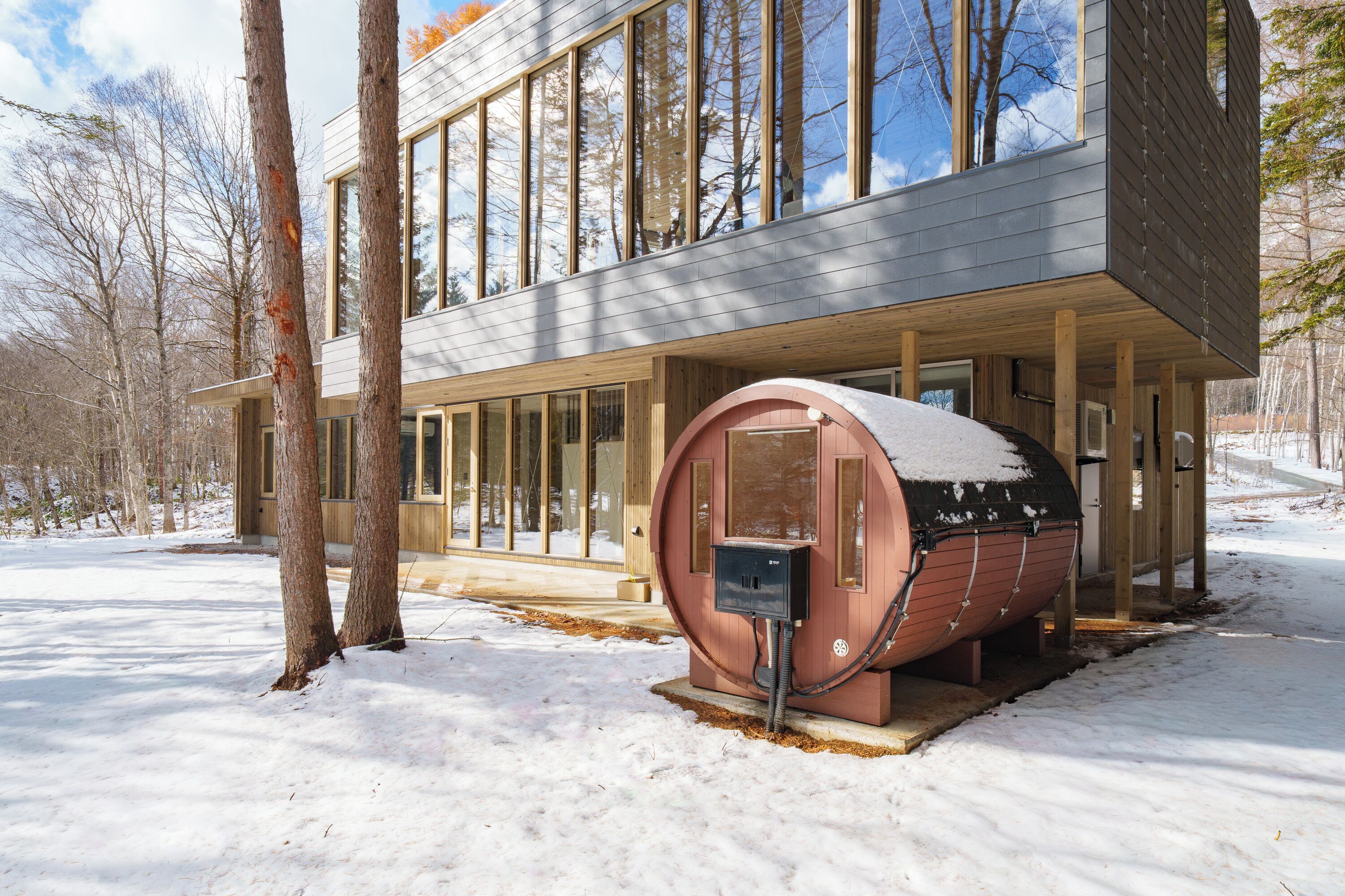 A modern house with large windows, a wooden deck, and a snow-covered barrel.