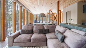 Un intérieur moderne avec un canapé d’angle, un plafond en bois, de grandes fenêtres et un coin repas.