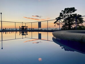 Une piscine entourée d’une clôture, avec un banc et une chaise, dont l’eau reflète les couleurs du coucher de soleil.