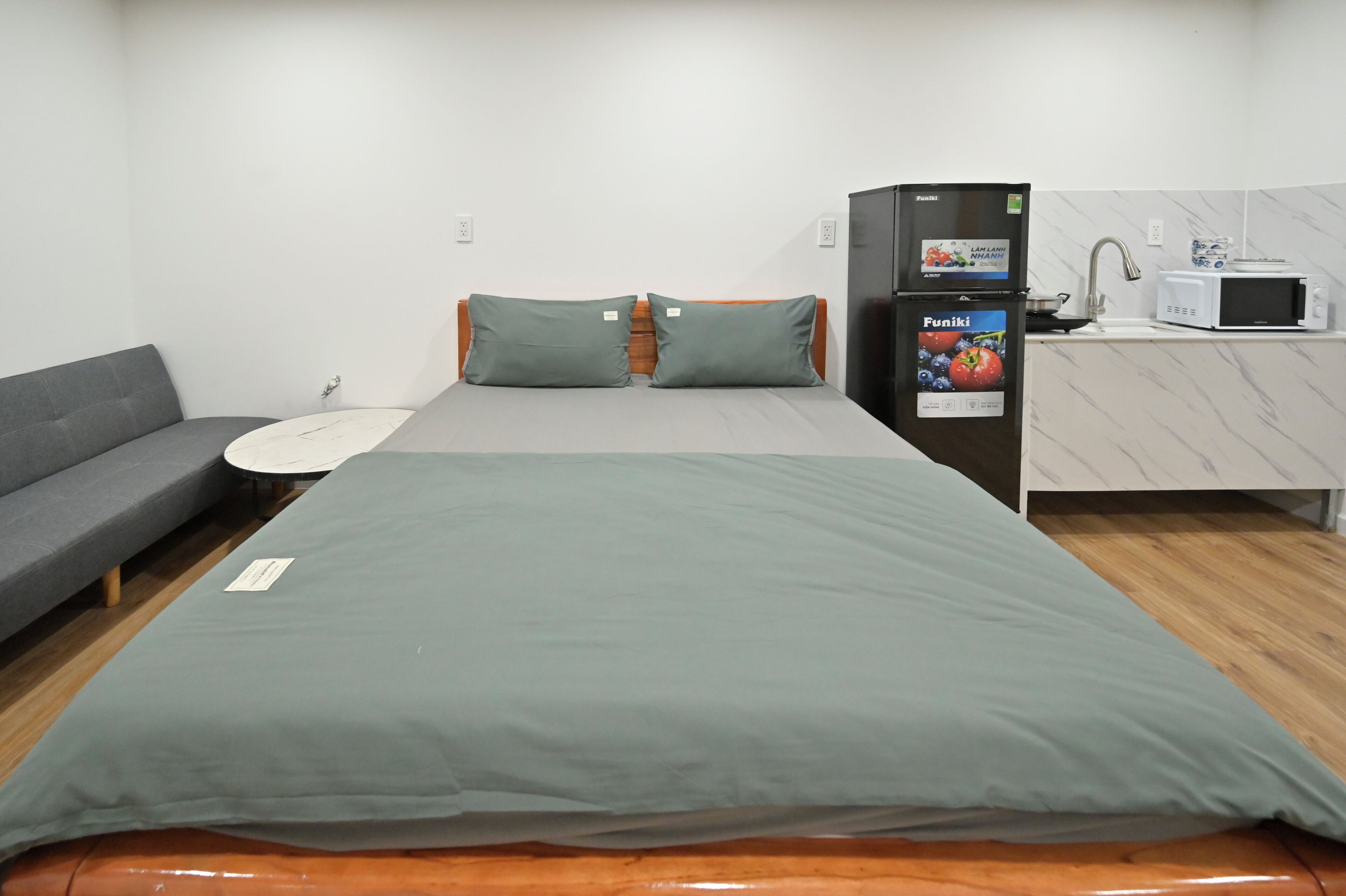A compact room with a bed, a gray sofa, a kitchenette with a microwave, and a refrigerator.