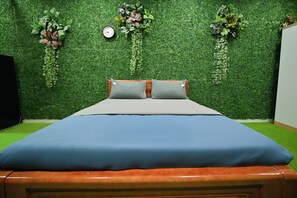 Una cama con colchón azul y dos almohadas, cabecera de madera, y pared de césped artificial verde con plantas colgantes y un reloj.