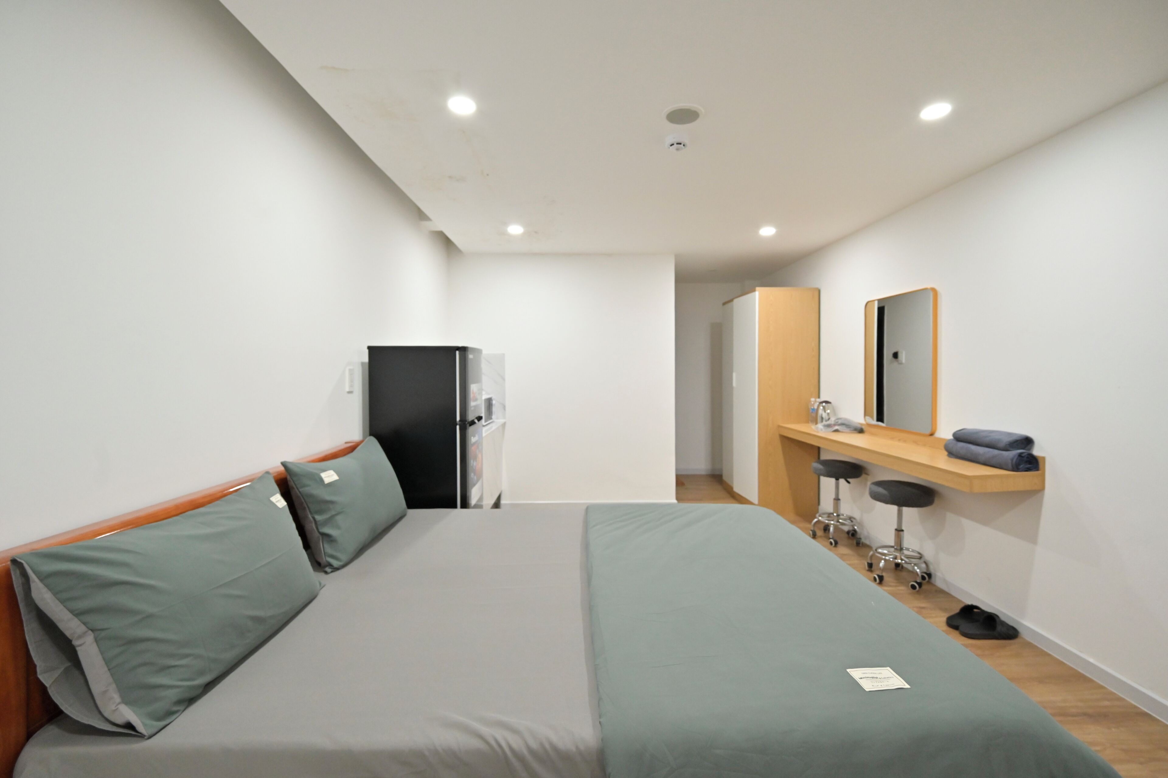 A hotel room with a bed, a desk with a mirror, a refrigerator, and a stool.