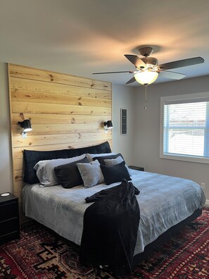 Un dormitorio con cabecera de madera, una cama con muchos almohadones, un ventilador de techo, una mesita de noche y una ventana con persianas.