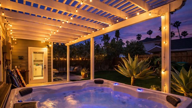 A hot tub under a covered patio with string lights, surrounded by a wooden fence and palm trees. - Block to Beach Family Home w/ King Bed, Jacuzzi & BBQ (San Diego)
