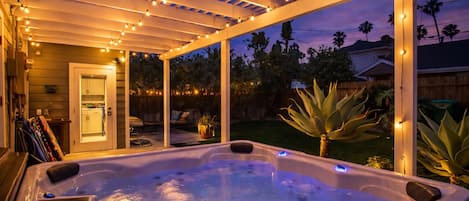 A hot tub under a covered patio with string lights, surrounded by a wooden fence and palm trees.