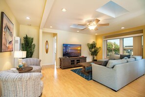 A living room with a sofa, armchairs, a TV, a ceiling fan, and a large window. - Block to Beach Family Home w/ King Bed, Jacuzzi & BBQ (San Diego)
