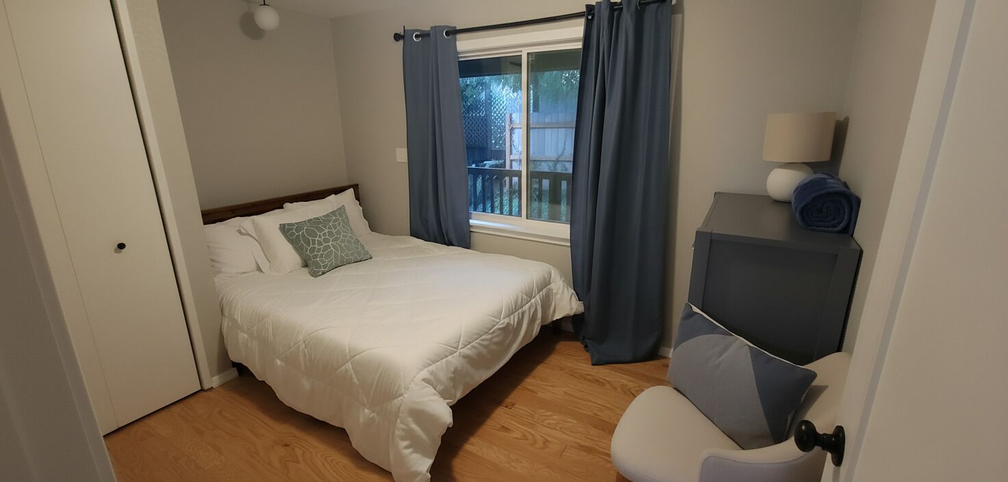 A bedroom with a bed, a window with curtains, a bedside table with a lamp, and a chair.