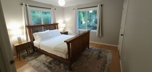 A bedroom with a wooden bed, two bedside tables with lamps, a large window with curtains, and a view of greenery outside.