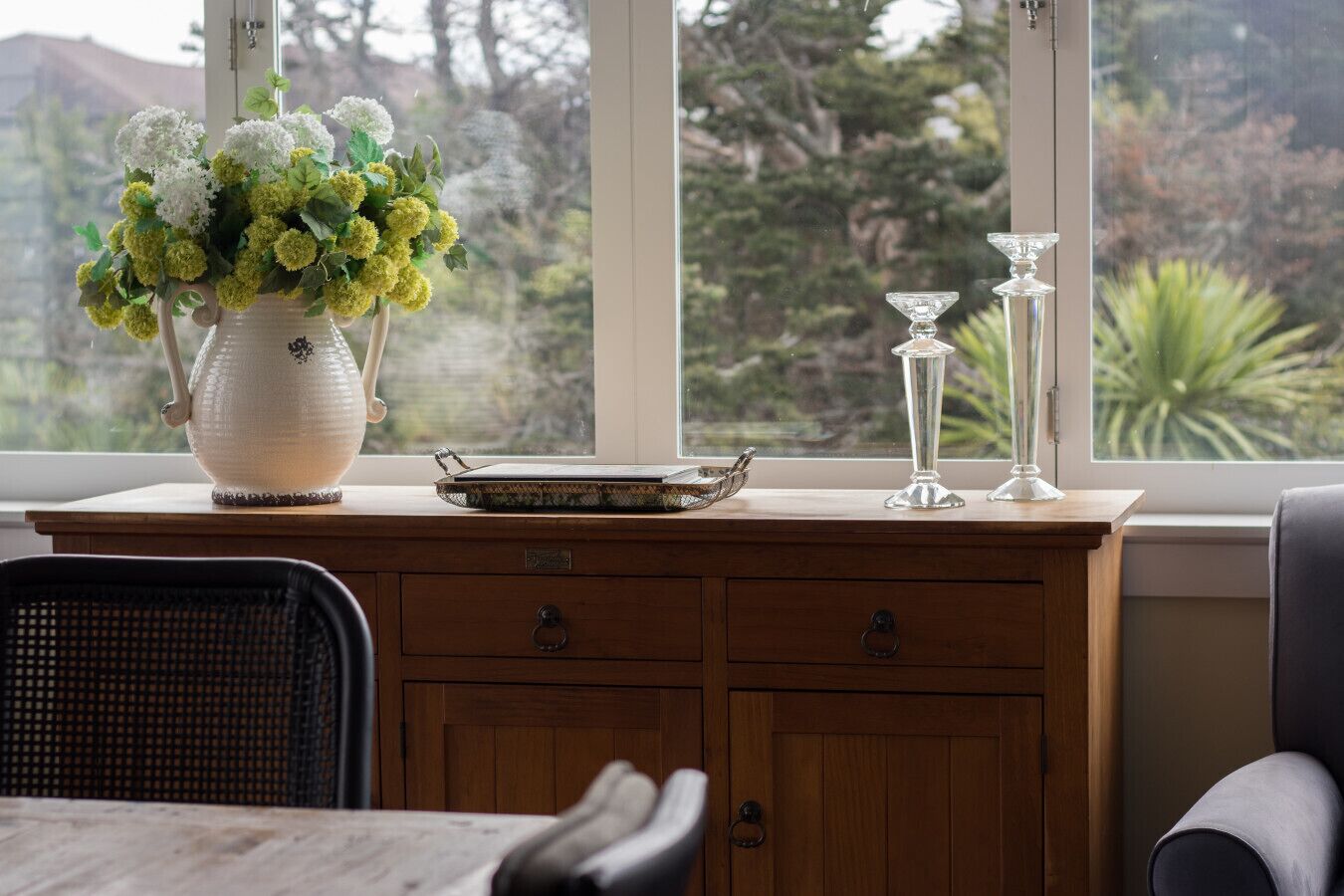 A wooden cabinet with a vase of flowers, a tray, and two glass candle holders on a windowsill.