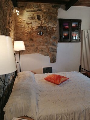 Ein Schlafzimmer mit einer Steinsäule, einem Bett mit einem gemusterten Überwurf, einem roten Kissen, einer Lampe und einem Fenster mit Blick.