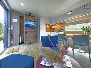 A modern hotel room with a TV, seating area, kitchenette, and a view of greenery.