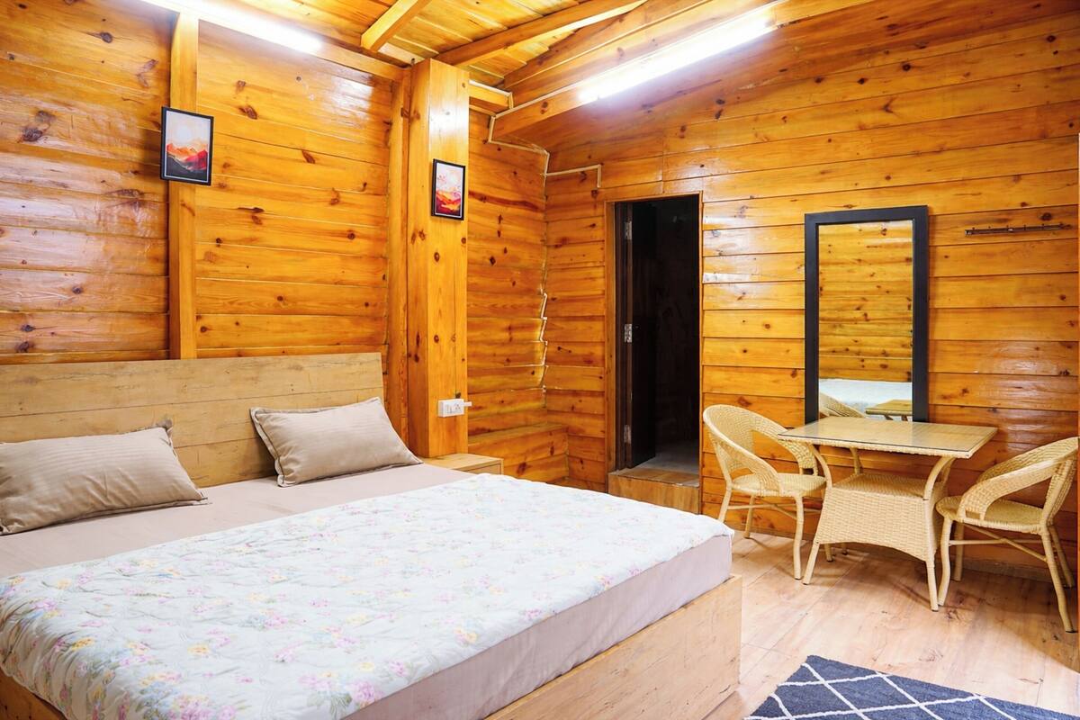 A wooden cabin room with a bed, two chairs, a table, and a mirror.