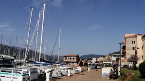 Una marina con diverse barche a vela ormeggiate, un percorso pedonale lastricato e edifici sullo sfondo.