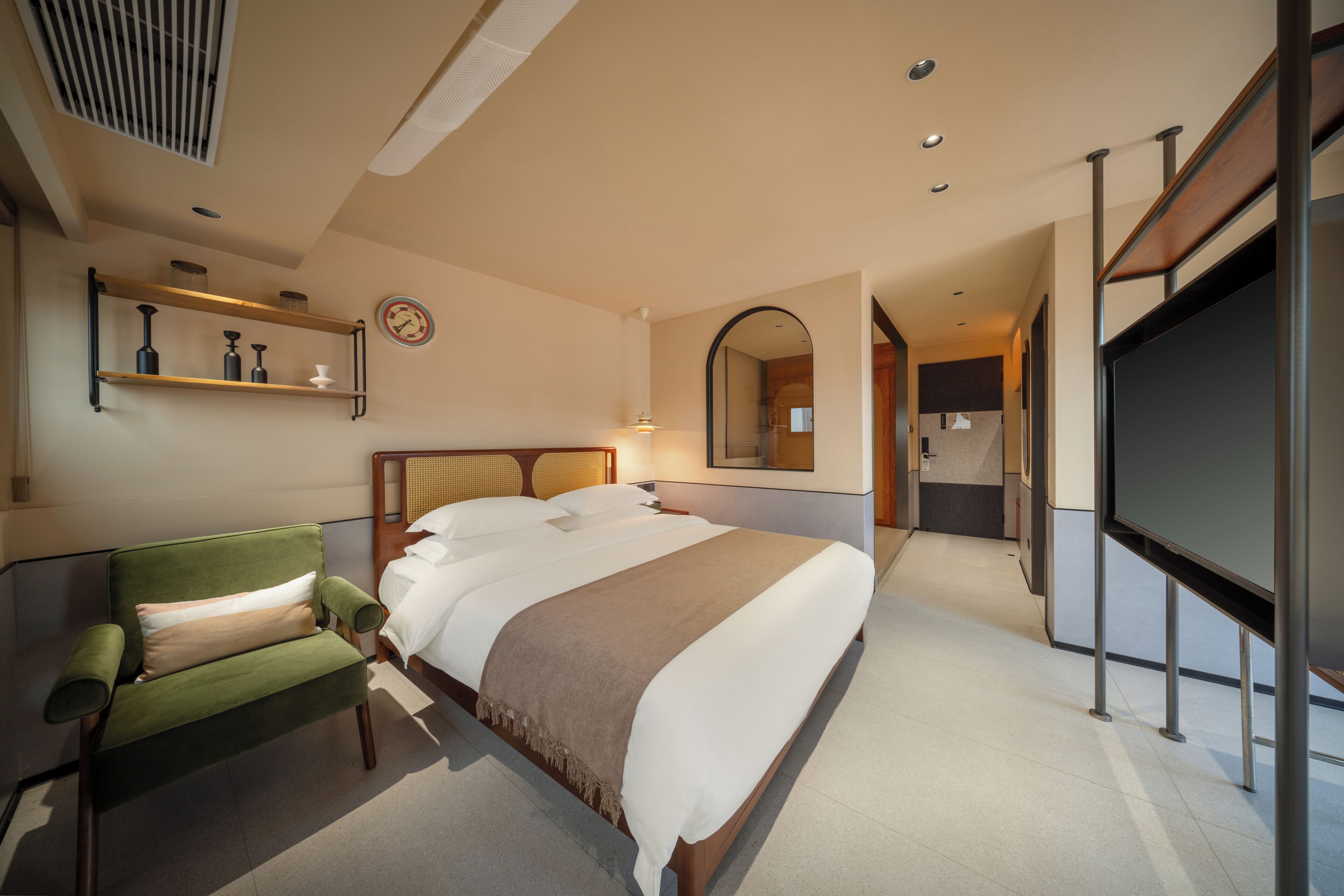 Una camera d'hotel con un letto, una poltrona verde, una TV, uno specchio e uno scaffale con oggetti decorativi.