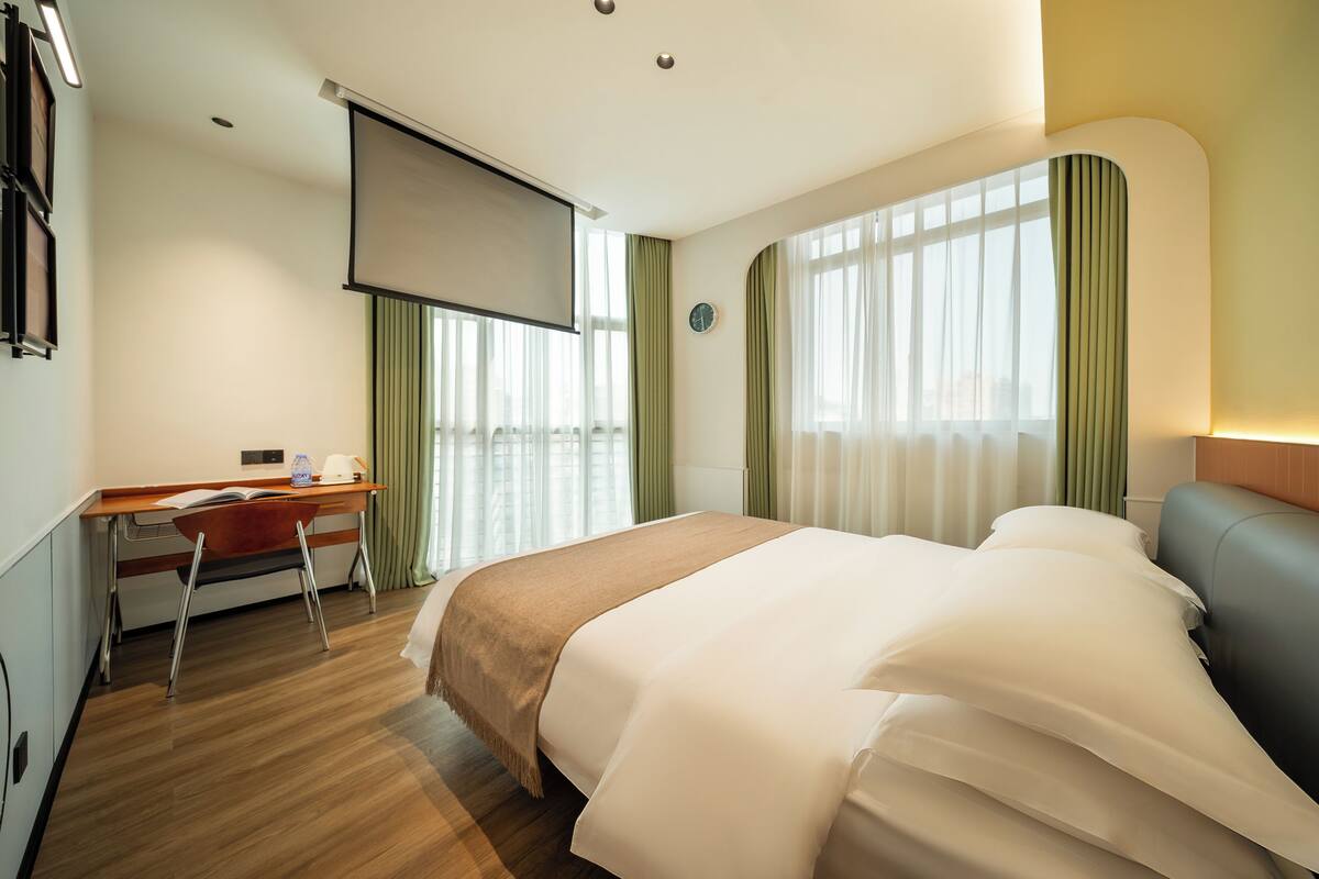 Habitación de hotel con cama, escritorio y un amplio ventanal con cortinas.