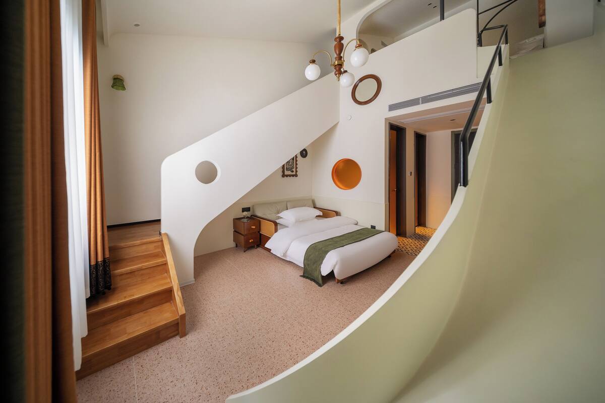Un dormitorio con cama, mesita de noche, cómoda, escalera y un ventanal pequeño.