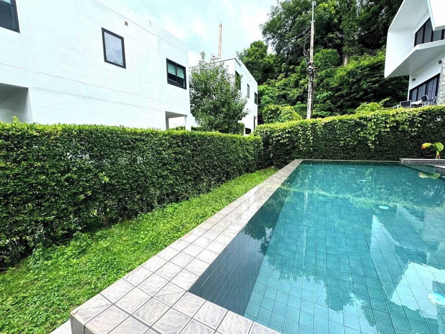 Piscina retangular com bordas azulejadas, cercada por um hedge bem aparado e um edifício branco com várias janelas.