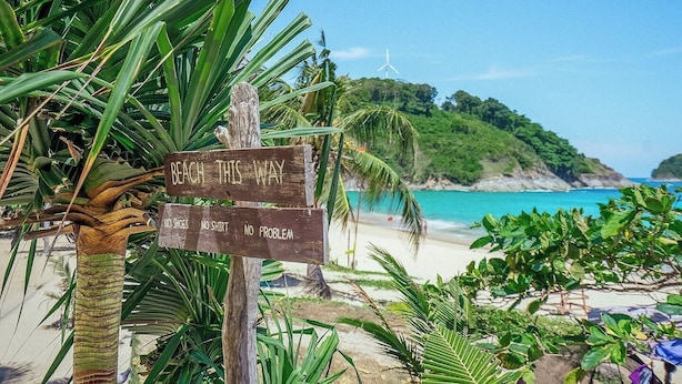 A wooden signpost with 'BEACH THIS WAY' and 'NO SHOES NO SHIRT NO PROBLEM' written on it, a beach, and a wind turbine in the background.