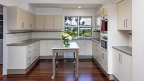 A modern kitchen with white cabinetry, a central island with a vase of flowers, a window with a view, and a built-in oven. - Large family home in a quiet rural location. (Isis Central)