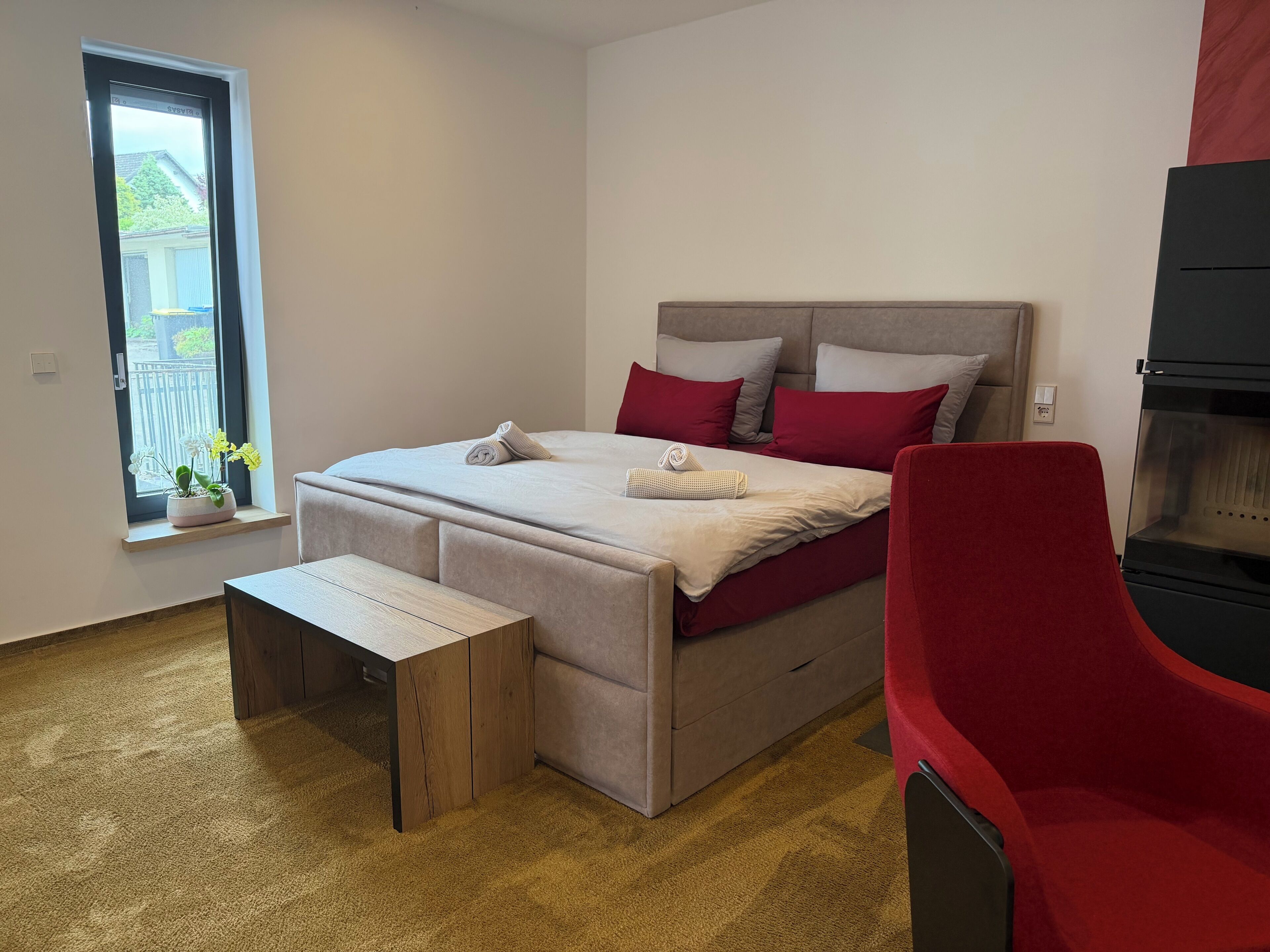 Ein Schlafzimmer mit einem Bett, einer Bank, einem roten Sessel, einem Fenster mit Pflanzen und einem schwarzen Gerät.