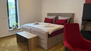 Ein Schlafzimmer mit einem Bett, einer Bank, einem roten Sessel, einem Fenster mit Pflanzen und einem schwarzen Gerät.