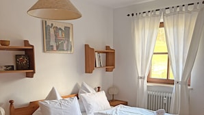 Un lit en bois avec du linge de lit blanc, une table de chevet, une lampe, une étagère avec des livres et un tableau au mur.