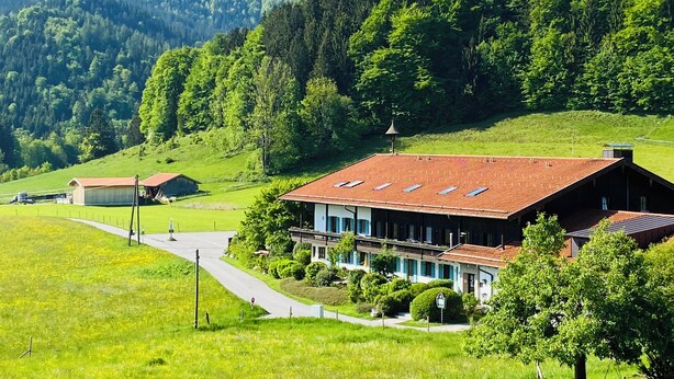 Un bâtiment rural avec un toit rouge, entouré de champs verdoyants et avec une chaîne de montagnes en arrière-plan.
