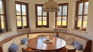 Une pièce avec une table ronde en bois, entourée de bancs ornés de motifs floraux, de grandes fenêtres à cadre en bois et un luminaire suspendu.