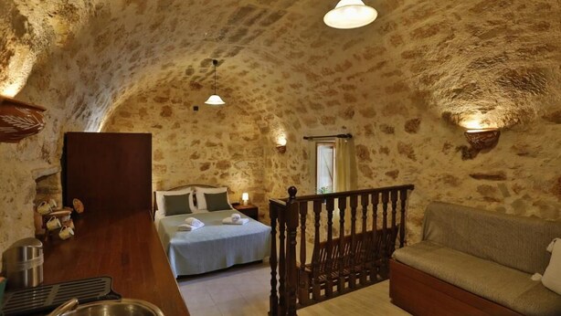 A rustic room with a stone wall, a bed, a wooden bench, a sofa, and a bar area with a sink. - Tholos by Praxithea Stone House & Pool (Chania)