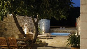 An outdoor patio area with stone flooring, wooden furniture, a pool, and a stone wall. - Tholos by Praxithea Stone House & Pool (Chania)